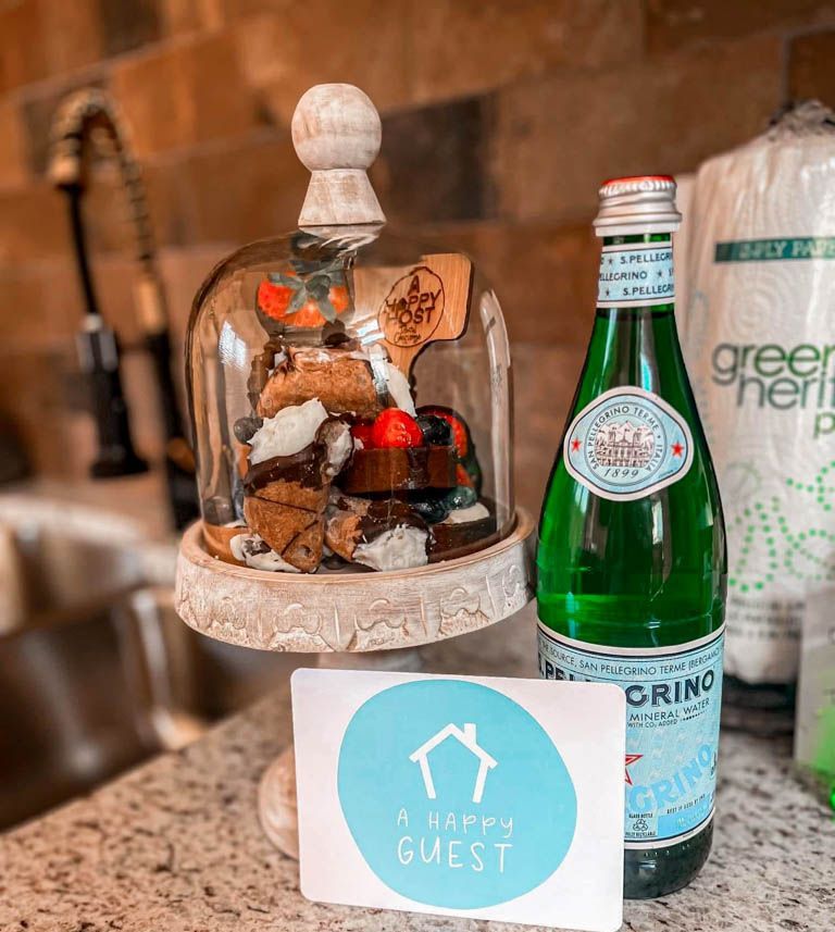 A Happy Guest welcome treats under a cloche with sparkling water at a rental cabin